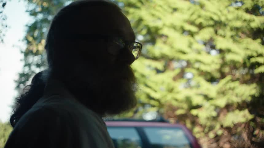 A bearded man with a ponytail sits on the porch talking to someone