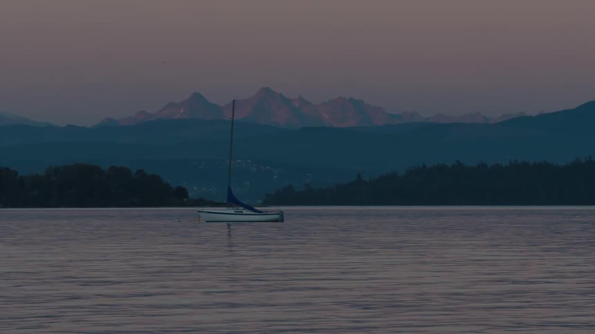 A snowcapped mountain stands tall against a soft pink sky reflected in the calm waters of a lake where a small sailboat glides peacefully