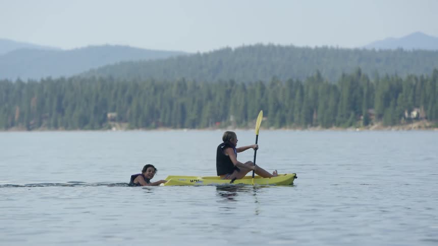 A kid paddling a board is towing another kid behind him