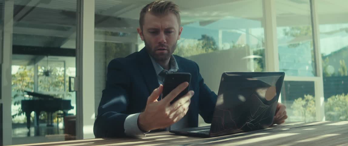 A young man in a black suit is sitting on his laptop and then takes his cell phone out of his jacket and then smiles