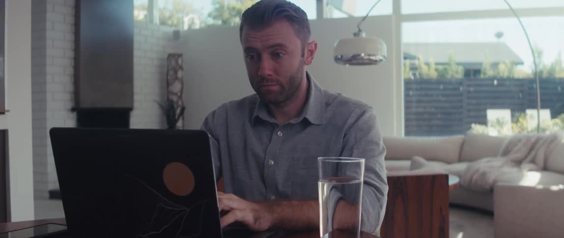 A man sitting is typing on laptop because it is in front of him next to a glass of water and he is sighing