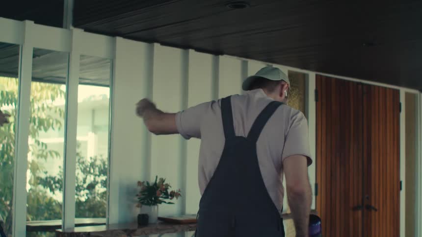 A man in a cap and overalls is cleaning a floor with a broom and he is dancing happily