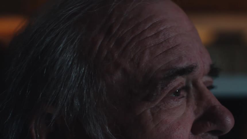 Close up of the face of an elderly man sitting staring straight ahead with lights from a screen reflecting off of him