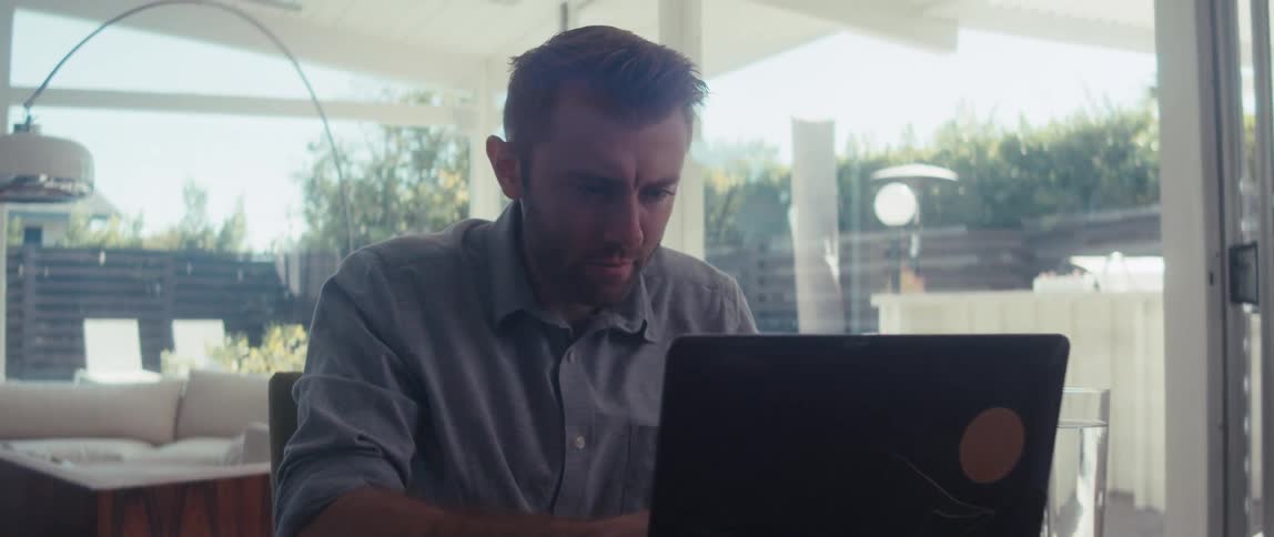 A young man is sitting and fiddling with a laptop in front of him and he puts his hand on his head in a nervous tone