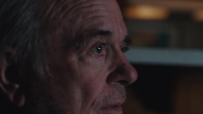 Elderly man sitting alone at home in a dimly lit room changes the television channel with a remote