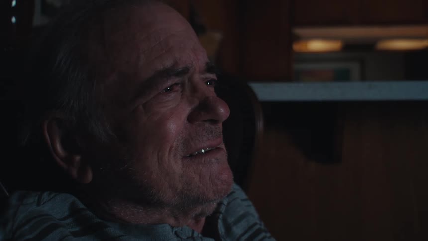 Elderly man frowns while watching television alone in a dimly lit area near the kitchen
