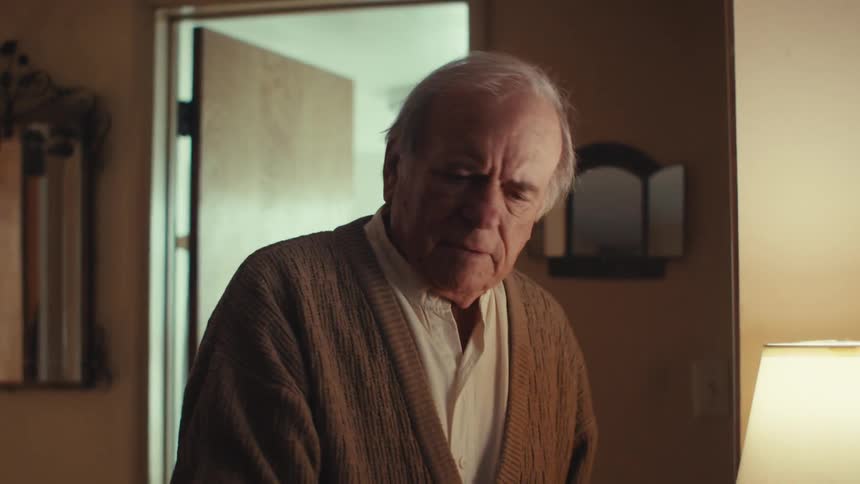 An elderly man sitting on a bed stands up to answer the phone at home