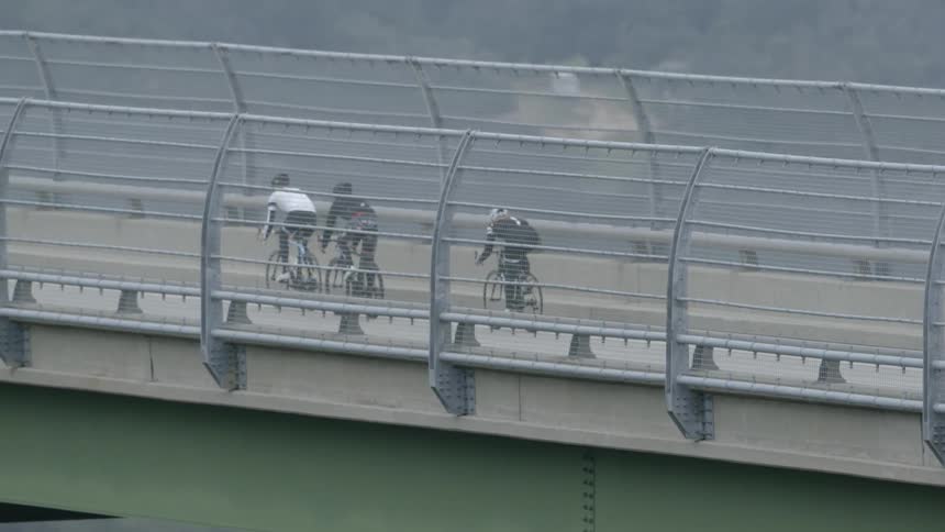 Three bicyclists are riding over a bridge in the opposite direction of traffic