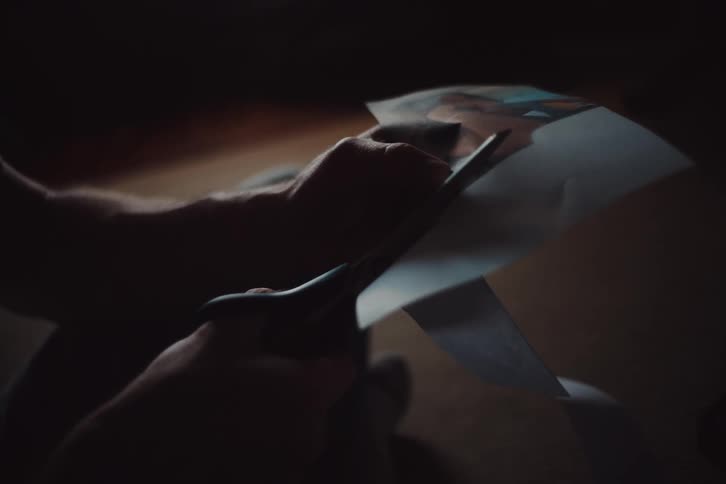 Man sitting in a dimly lit room cuts out a picture printed on a piece of paper