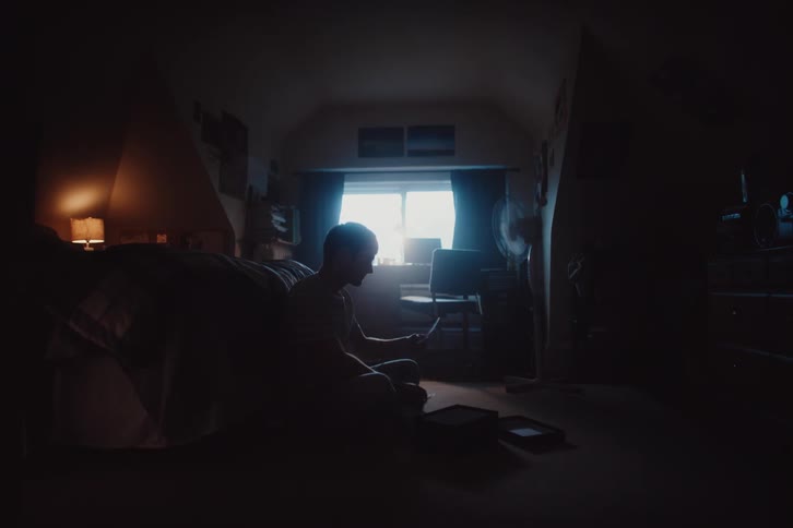 A person sits on the floor of a dimly lit room looking through a box of photos
