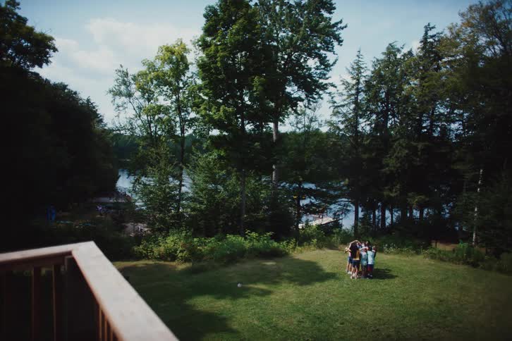A group of children stands in a circle on a grassy area with trees and a lake in the background