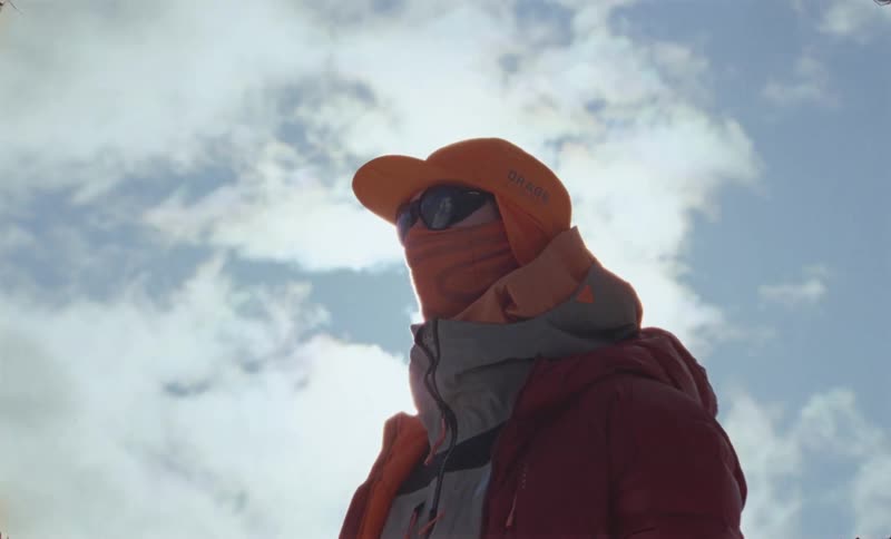 A climber bundled in warm gear and sunglasses looks upwards against a backdrop of a cloudy blue sky