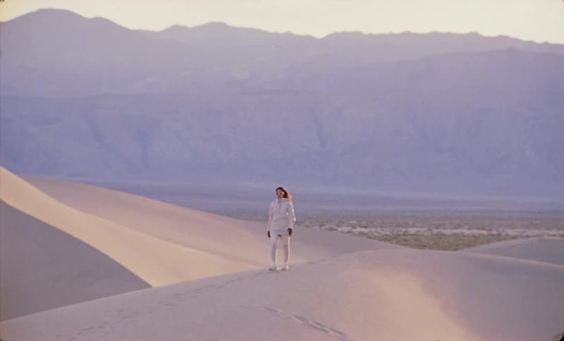 A solitary figure walks across sunlit sand dunes under a purpletinged mountain horizon