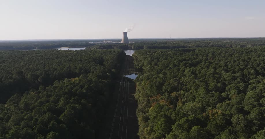 Power lines lead through the forest and over a river towards a smoking power plant on a slightly cloudy day