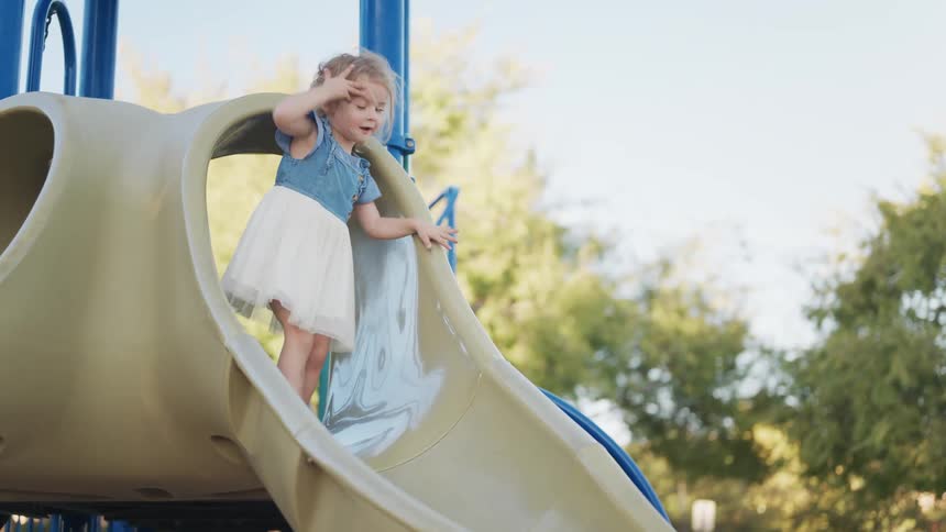 A little girl in a denim dress and tulle skirt climbs up and slides down a playground slide