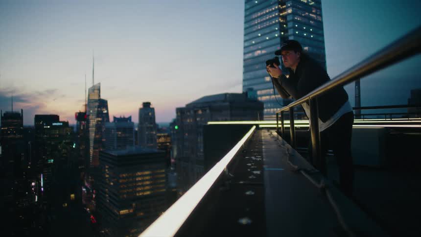 A man in a black cap is leaning on a railing at the top of a building using a camera to photograph the city