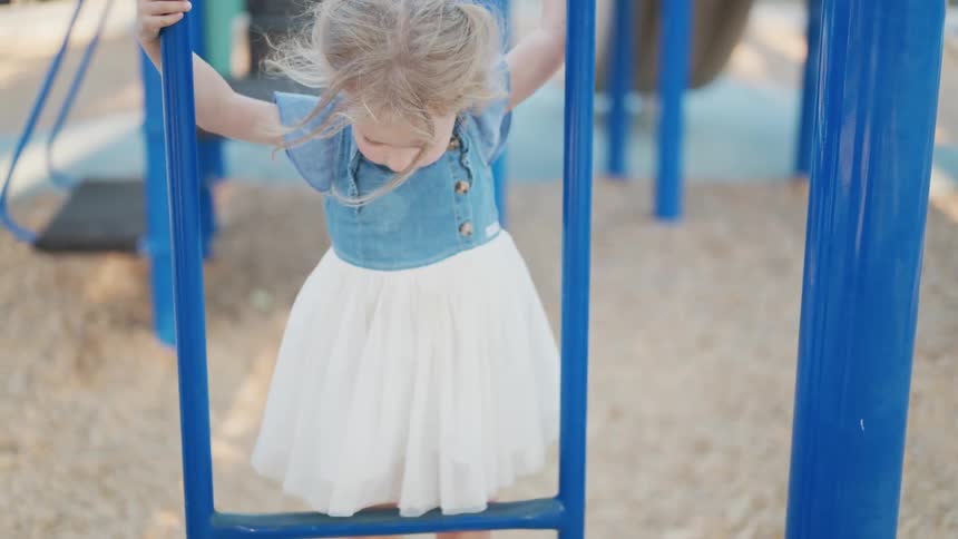 A young girl in a denim top and white dress plays on a blue metal playground structure