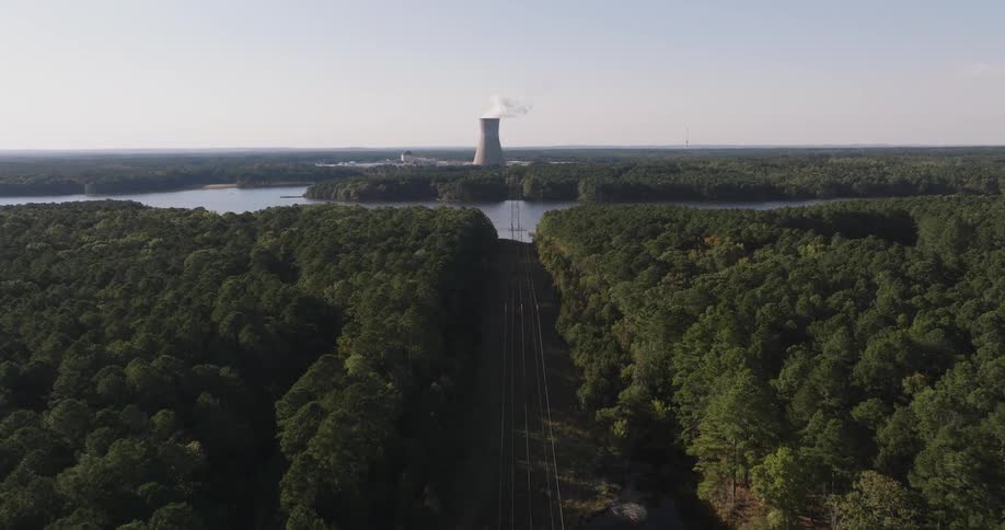 Aerial shot of power transmission towers in a forested area with a river and a factory in the background