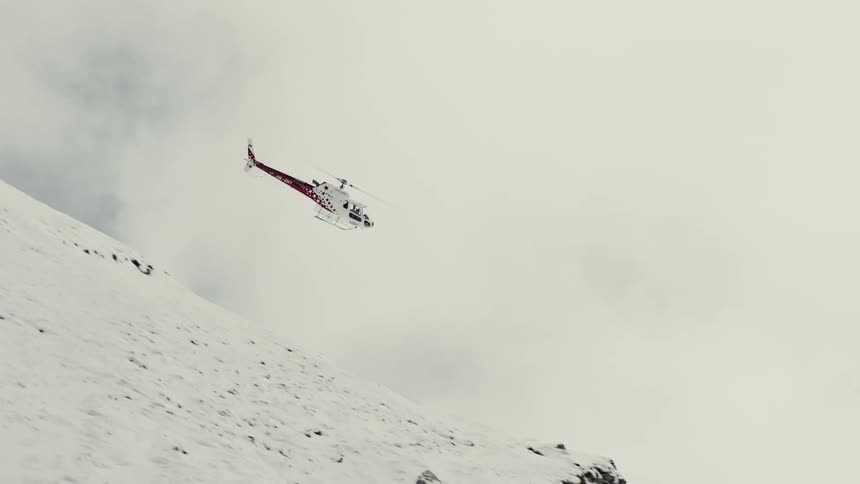A white helicopter with red accents flies over a snowcovered mountain peak