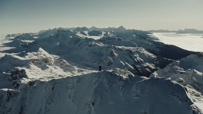 A solitary figure trekks a snowy mountain peak surrounded by breathtaking alpine vistas
