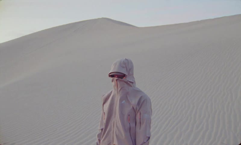 A person with his face covered by a jacket is in a desert region with a sand dune behind