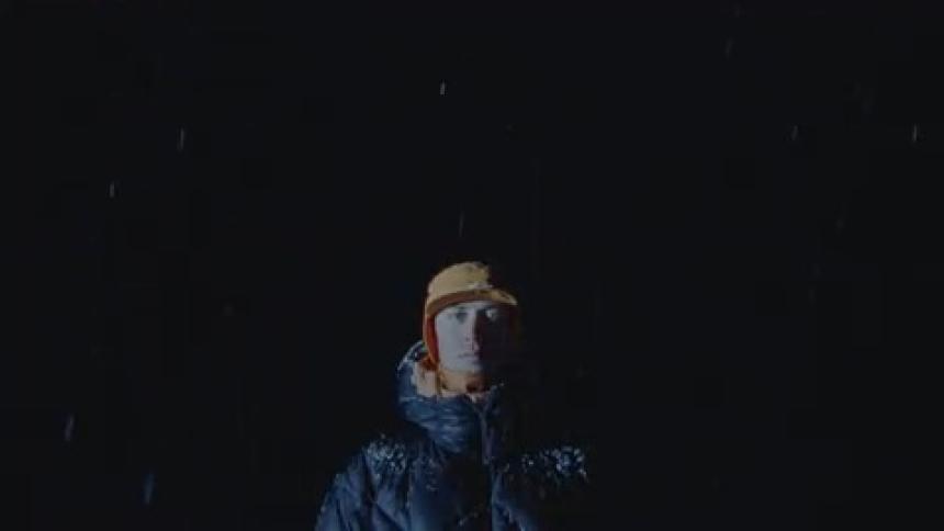 A man standing and wearing warm clothes is in a very dark environment that is snowing and he is staring straight ahead