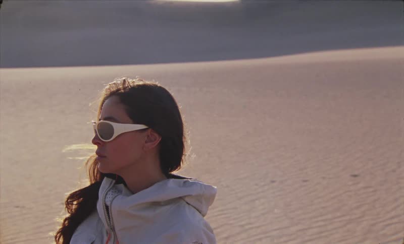 A young woman wearing white framed sunglasses and a jacket is standing in a desert looking to her right