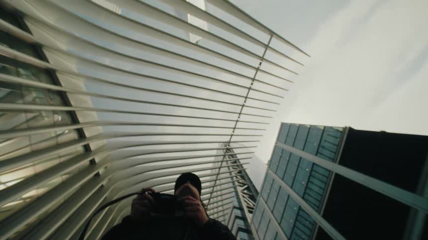 A person holding a camera walking under a modern architectural structure with tall buildings in the background