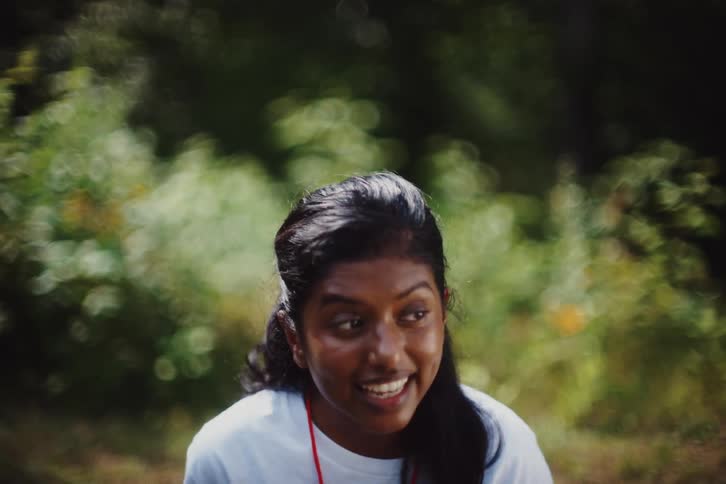 A cheerful woman is outdoors smiling brightly and waving her hands energetically with a lush green background adding to the vibrant and lively mood