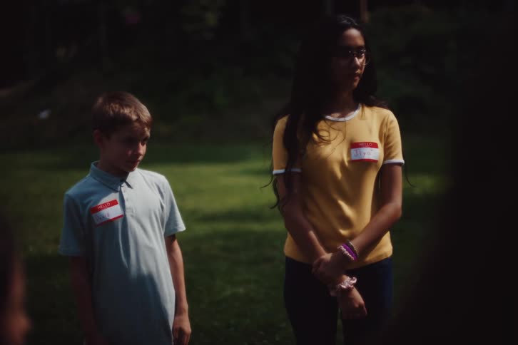 A group of children are standing on a lawn and there is a little boy who is moving nervously from side to side in a row everyone raises their right hand up