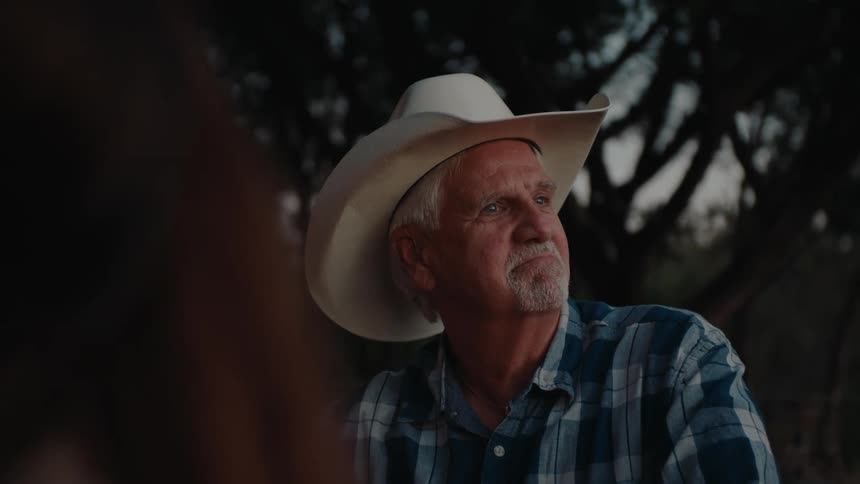 Elderly man wearing a white cowboy hat and blue plaid shirt, gazing thoughtfully into the distance during twilight, with dark silhouetted trees in the background.