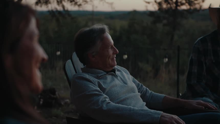 Middle-aged man reclining in an outdoor chair at dusk, smiling and engaged in conversation, with blurred greenery and warm twilight tones in the background.