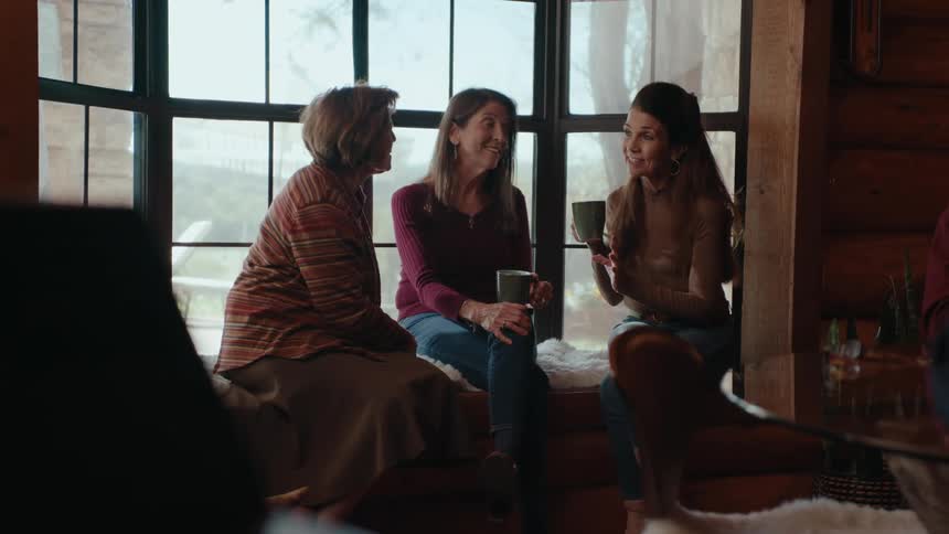 Three women are seated by a large window chatting and holding mugs in a rustic style room