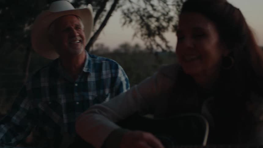 A woman plays the guitar while a man in a cowboy hat sings in a naturally dim setting