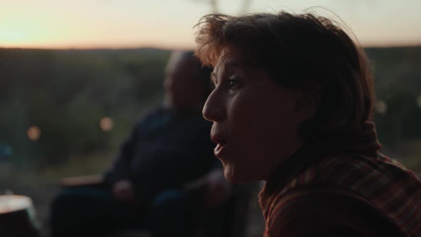 A seated woman starts laughing and then starts talking to someone in front of her and behind her there is a man who is also sitting