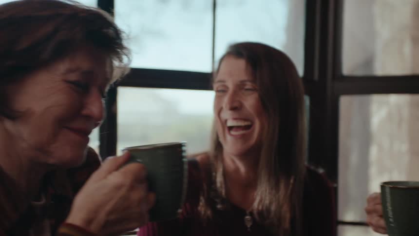 Three older woman sit inside a house holding coffee cups and laughing