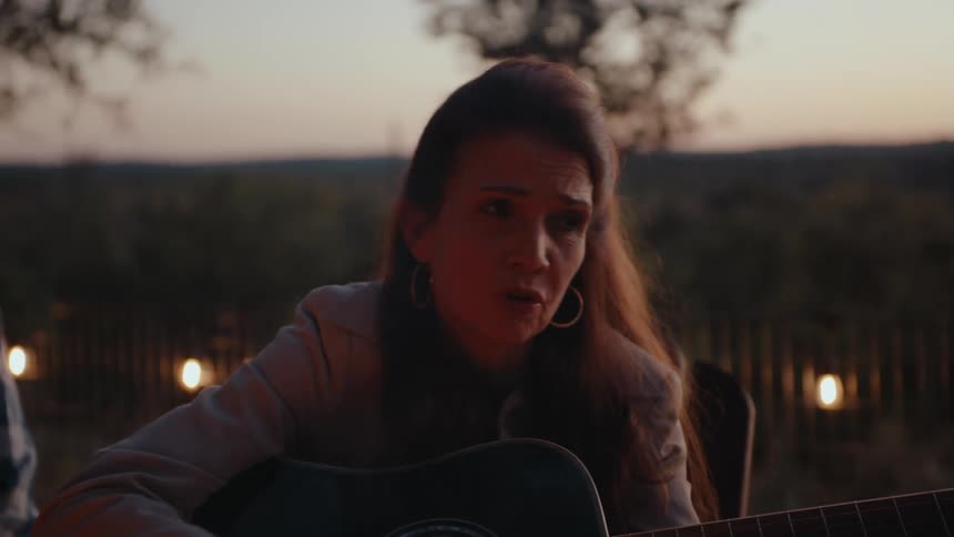 A woman sitting playing a black acoustic guitar near a smoking campfire
