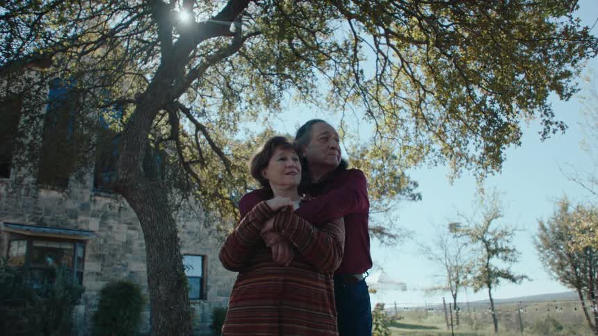 Two people stand under a large tree one embracing the other with a stone building and a clear sky