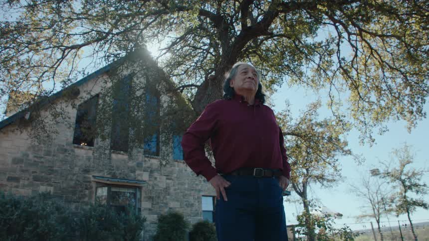 A person stands outdoors with hands on hips in front of a stone building with large windows under a tree with sunlight filtering through the branches