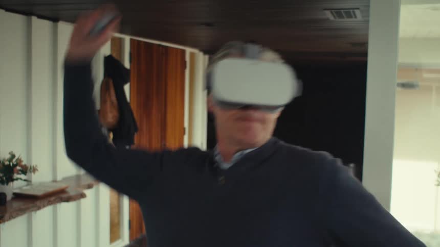 A grayhaired man wearing virtual reality glasses on his head and moving his arm forward and then he takes off the glasses and stares straight ahead