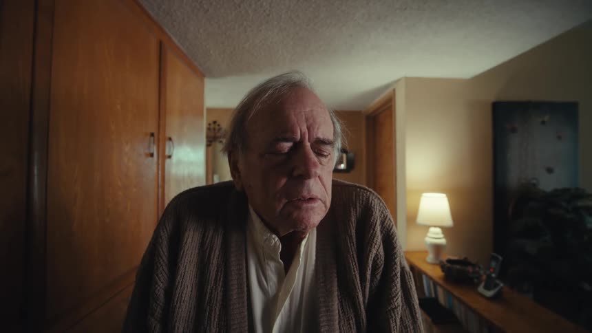 An elderly man wearing a sweater walks into a room looking confused