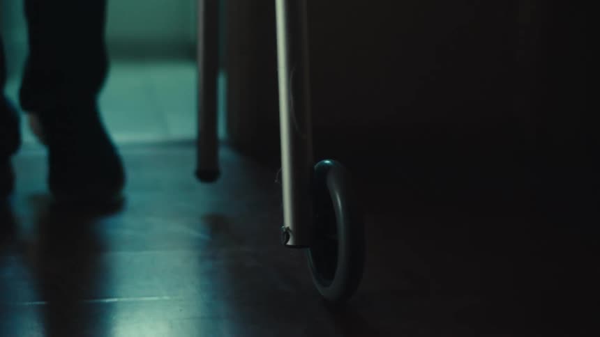 The feet of an elderly pushing a walker in a dimly lit room