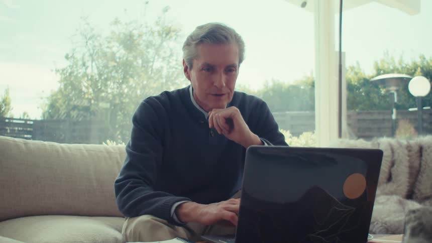 A grayhaired man is sitting on a sofa and fiddling with a laptop in front of him