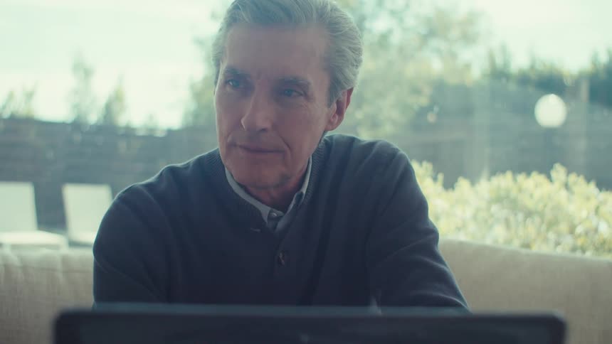 A grayhaired man is sitting on a couch and looking to the side then looking forward where there is a laptop