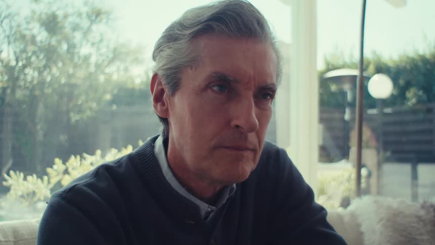 An man with gray hair is sitting indoors near a window with a garden visible outside