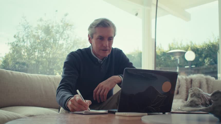 A man is sitting on a couch writing in a notebook with a pen and has a laptop open on a table in front of him