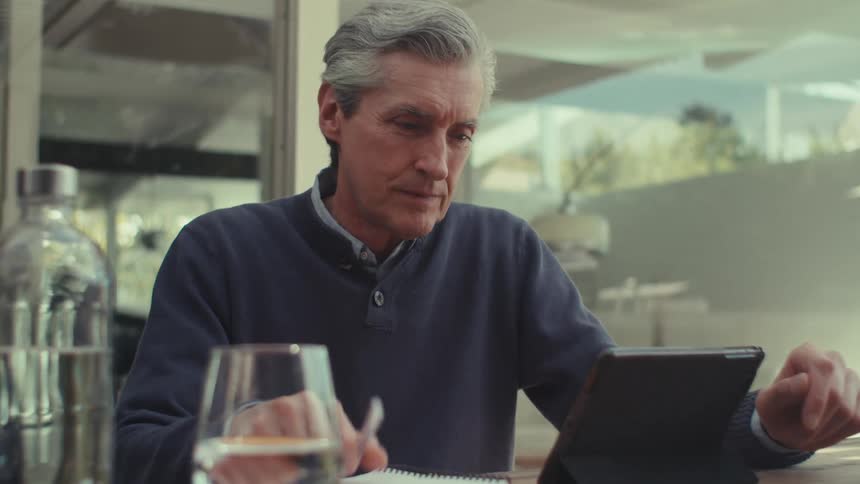 Older man takes notes while using a tablet at a table near the window