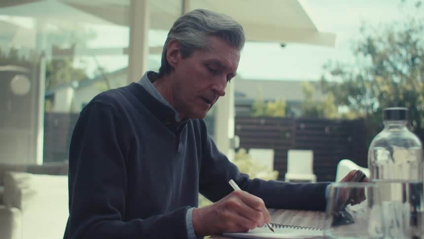 A grayhaired man is sitting while writing on a paper and touching the screen of a tablet with his other hand