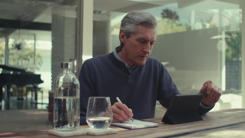 White man sitting down at a table outdoors writing on a notebook while looking at a screen in a contemplative way during daytime