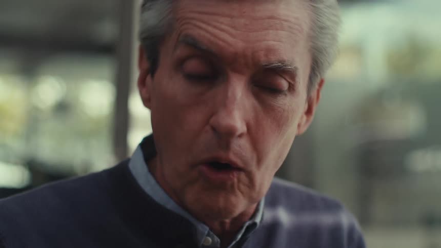 A person with gray hair wearing a dark sweater over a collared shirt talking outdoors